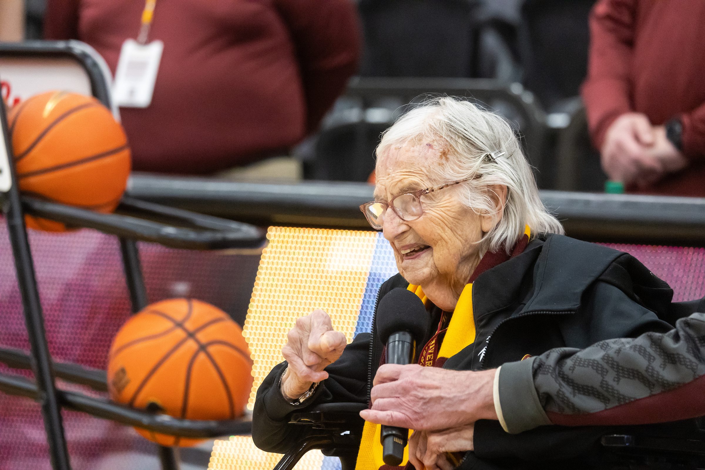 Sister Jean Dolores Schmidt, BVM, speaks before the Ramblers took on Fordham on Jan. 22, 2025. A woman using a wheelchair speaks into a microphone in front of a cart full of basketballs at a Loyola University Chicago basketball game