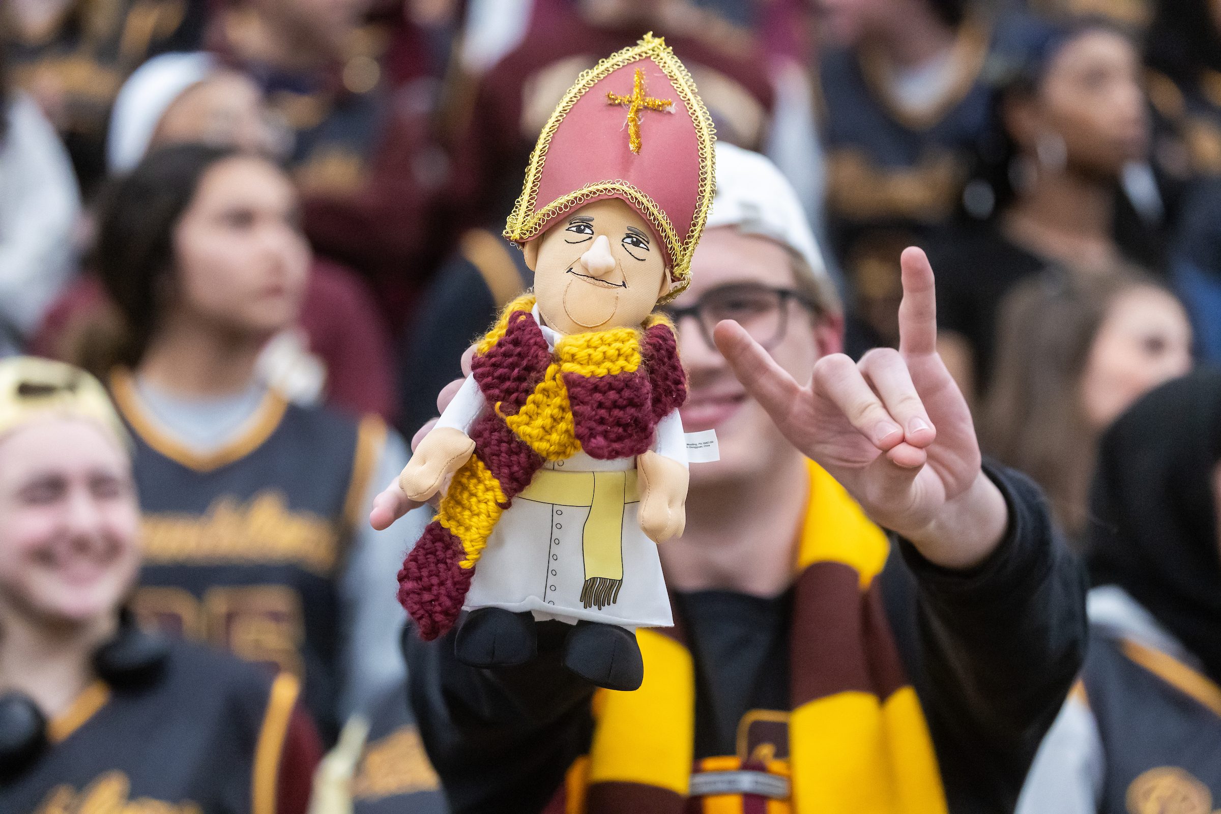 Students cheer on the Ramblers during a men's basketball game on Jan. 22. The Ramblers beat Fordham 70-66. A Loyola University Chicago student wearing a maroon and gold scarf holds up a Pope puppet and makes the wolf hand symbol with a crowd of students in the background