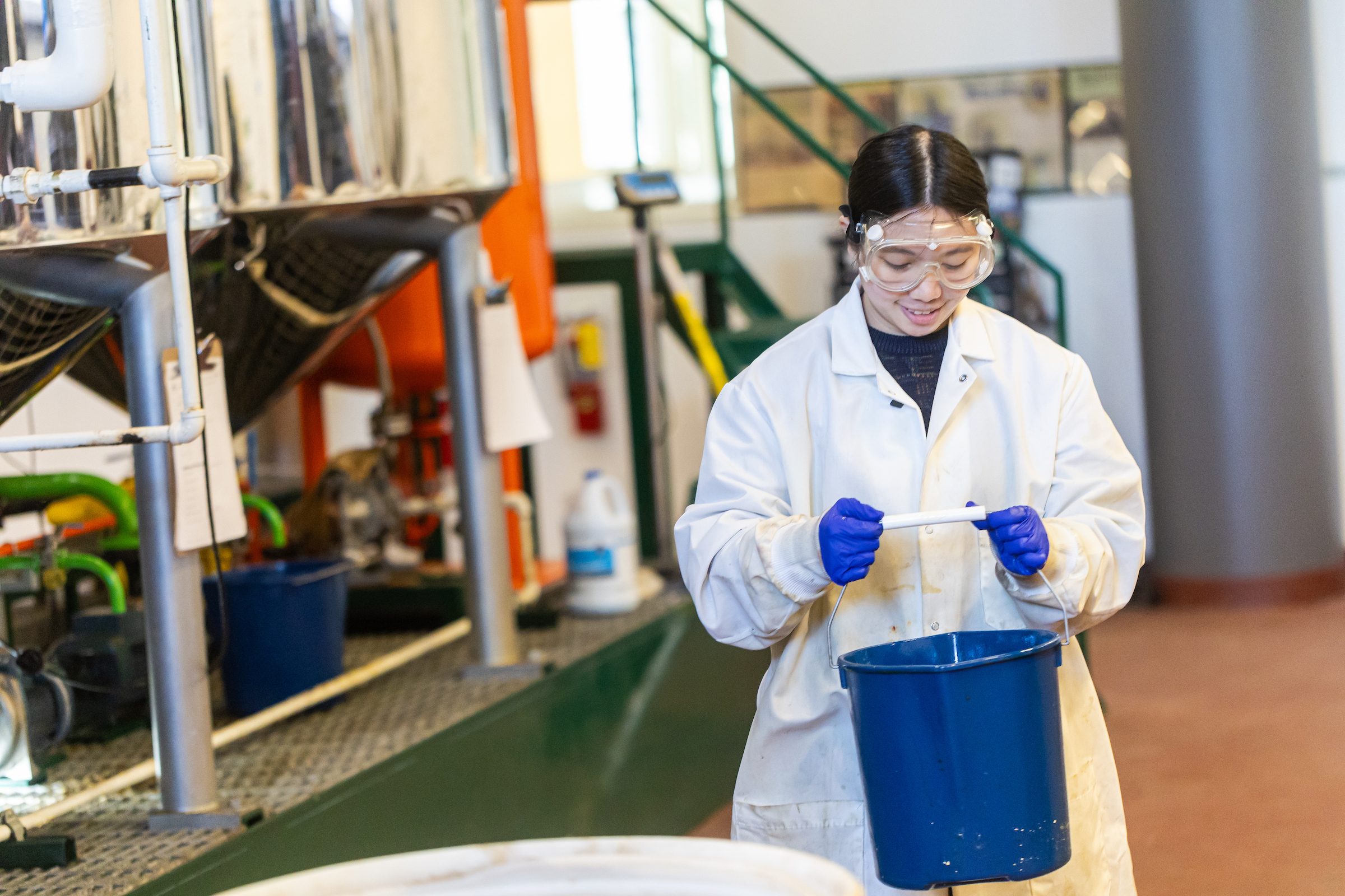 A Loyola University Chicago student wearing a lab coat and goggles carries a bucket in a lab