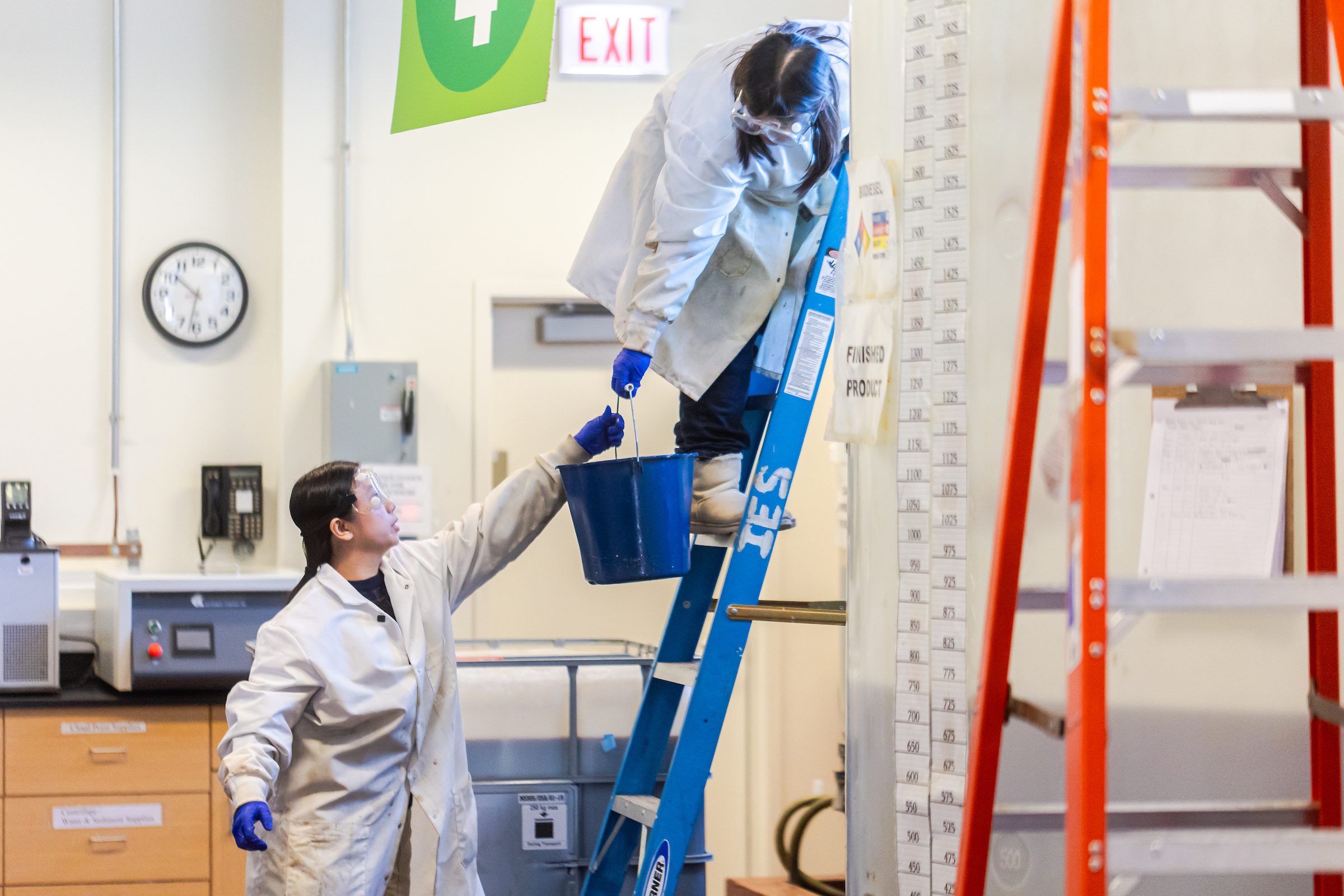 A Loyola University Chicago student wearing a lab coat passes a bucket to another sutdent who is standing on a ladder in a lab