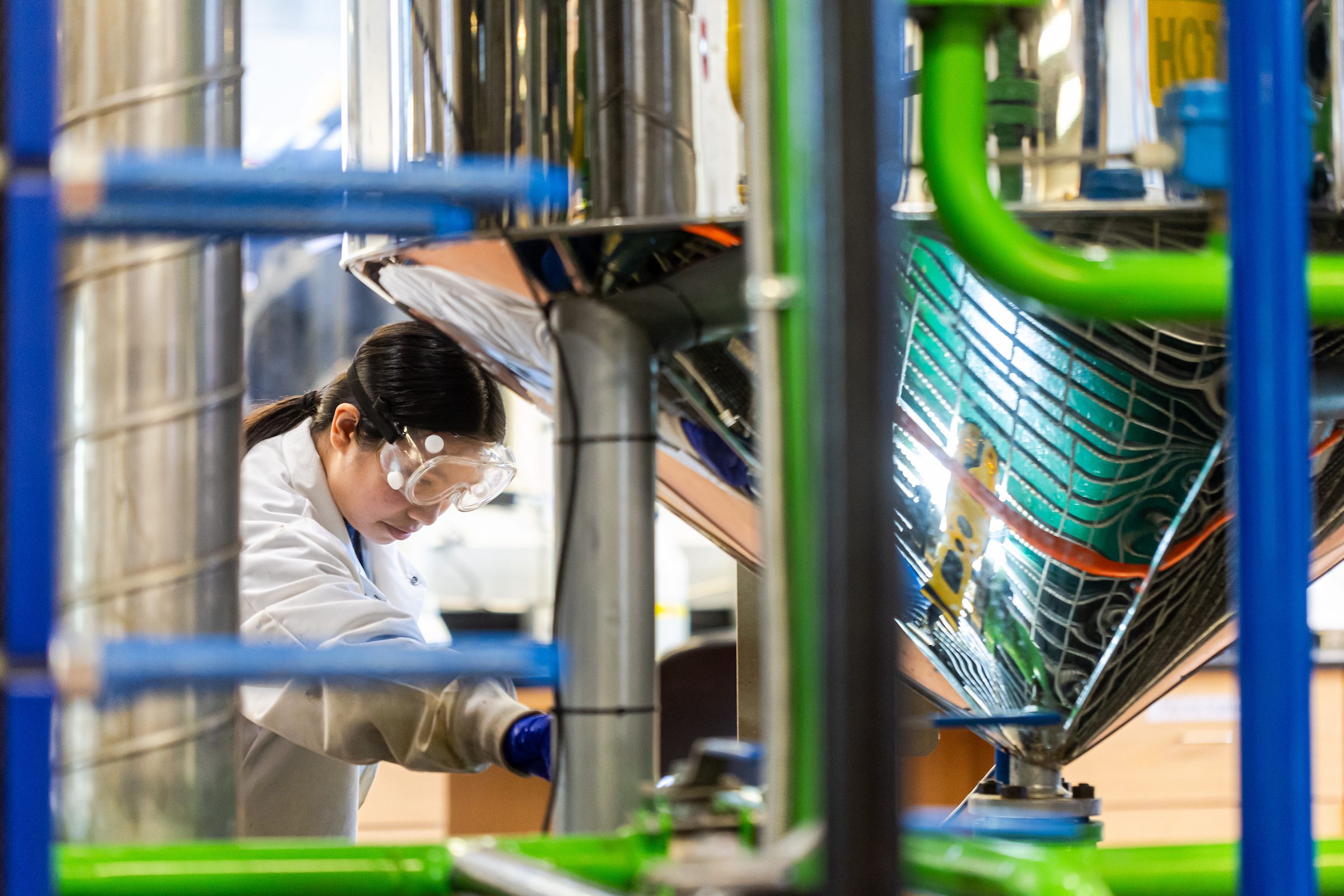 A Loyola University Chicago student wearing a lab coat and goggles works in a lab with pipes