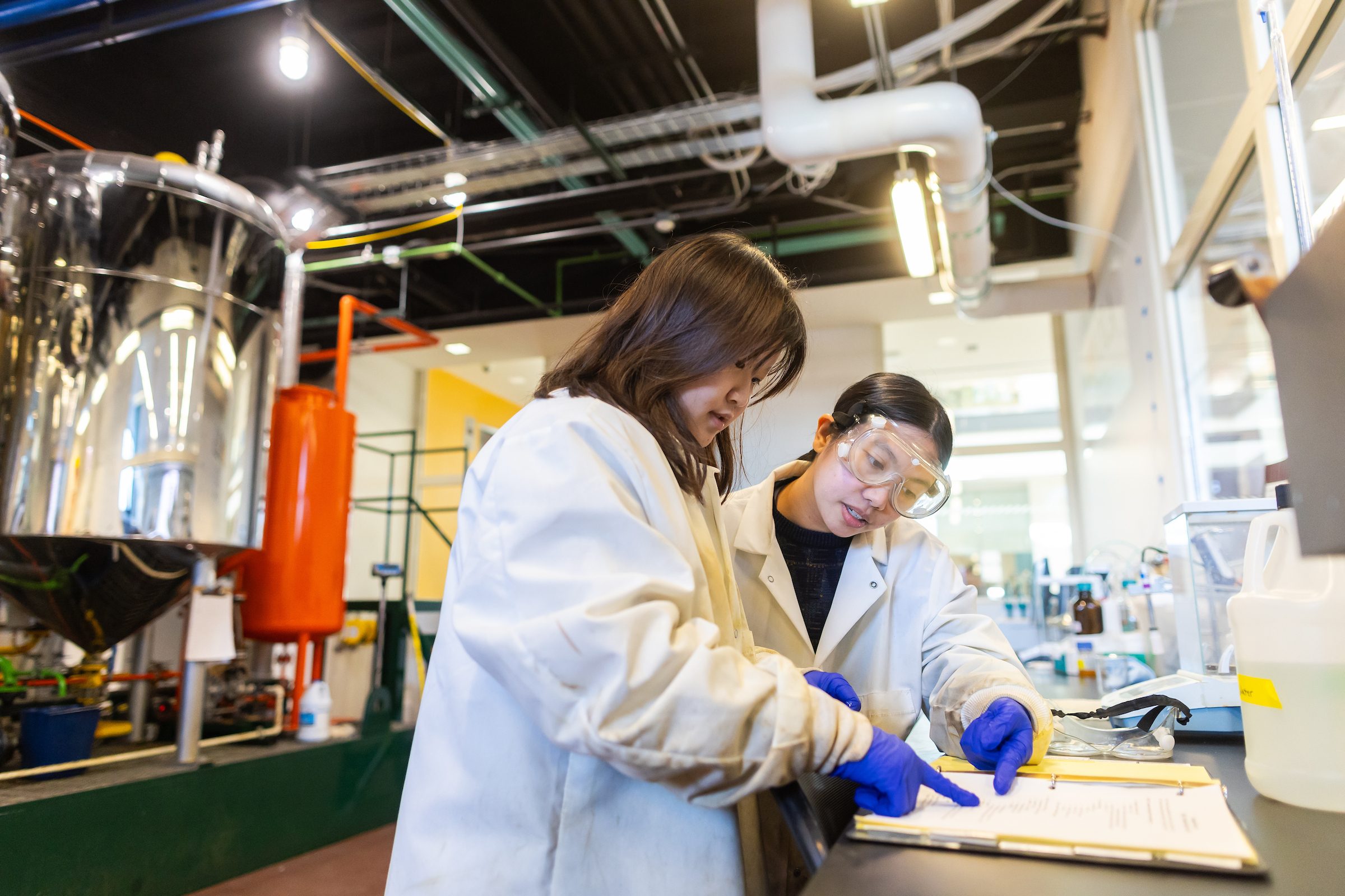 Two Loyola University Chicago students wearing lab coats and goggles point to a binder as they speak in a lab on campus