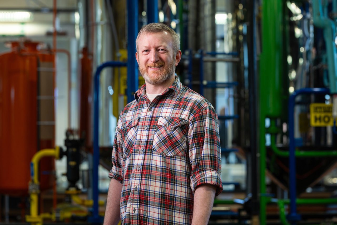 A man wearing a plaid flannel shirt poses for a portrait in a lab with multicolored pipes in the background