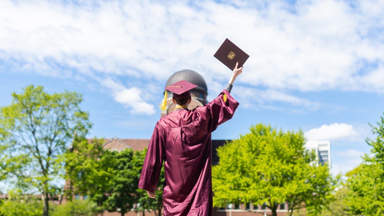 A Loyola University Chicago graduate wearing a cap and gown holds up their degree outside of Gentile Arena with trees in the background