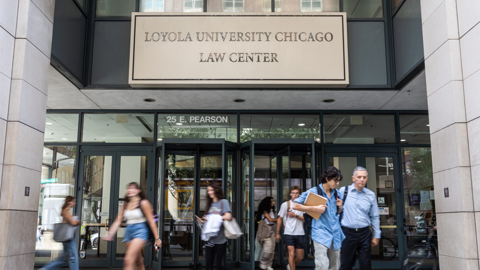 Students walk in and out of the doors of a building. A sign on the building reads 'Loyola University Chicago Law Center