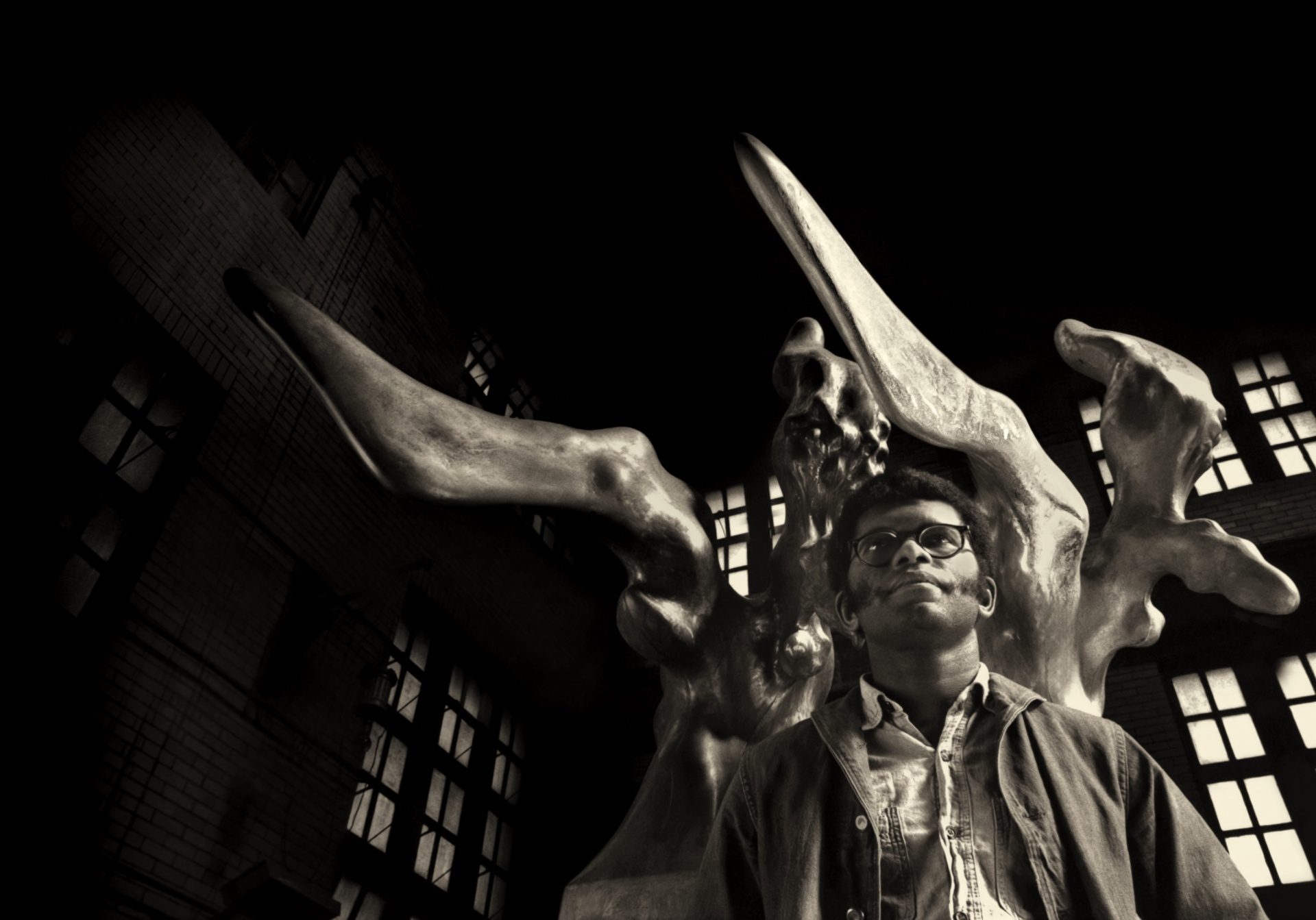 Young man stands in front of a large sculpture. Image is black and white.