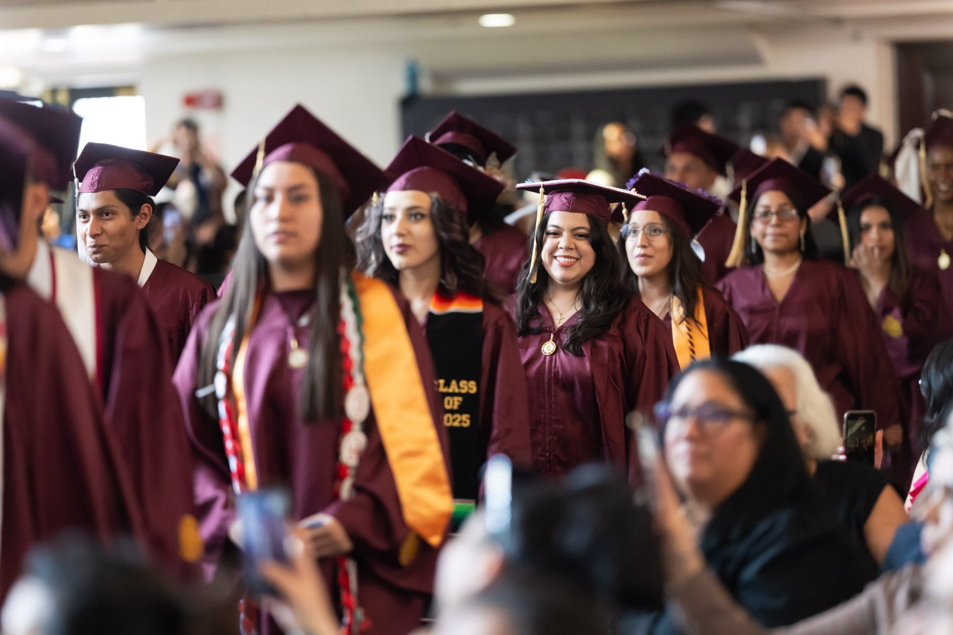 Loyola University Chicago students in maroon robes with gold tassels line up to receive their diplomas at an indoor commencement ceremony