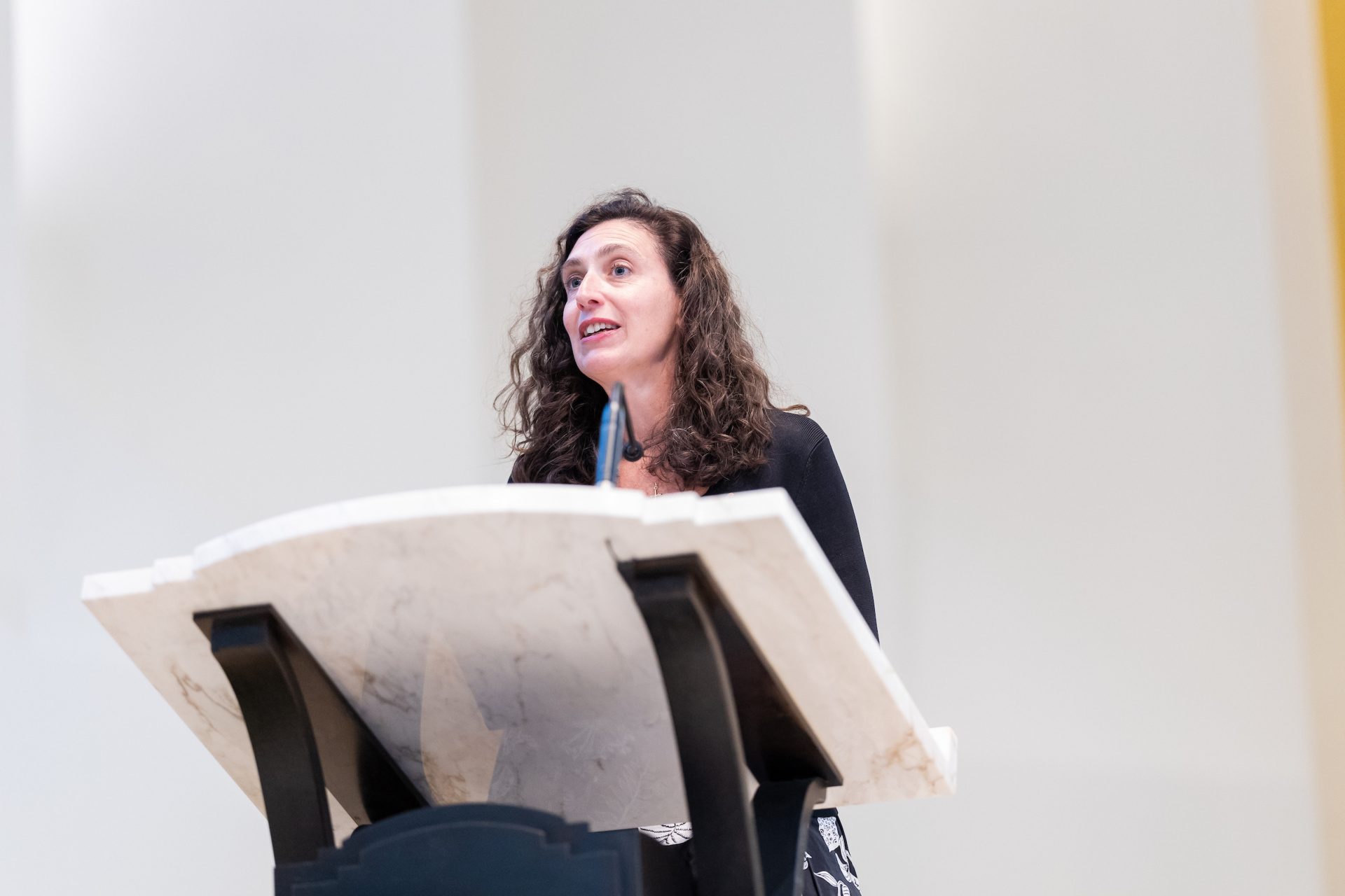 A woman speaks from a podium in a bright, well-lit room.
