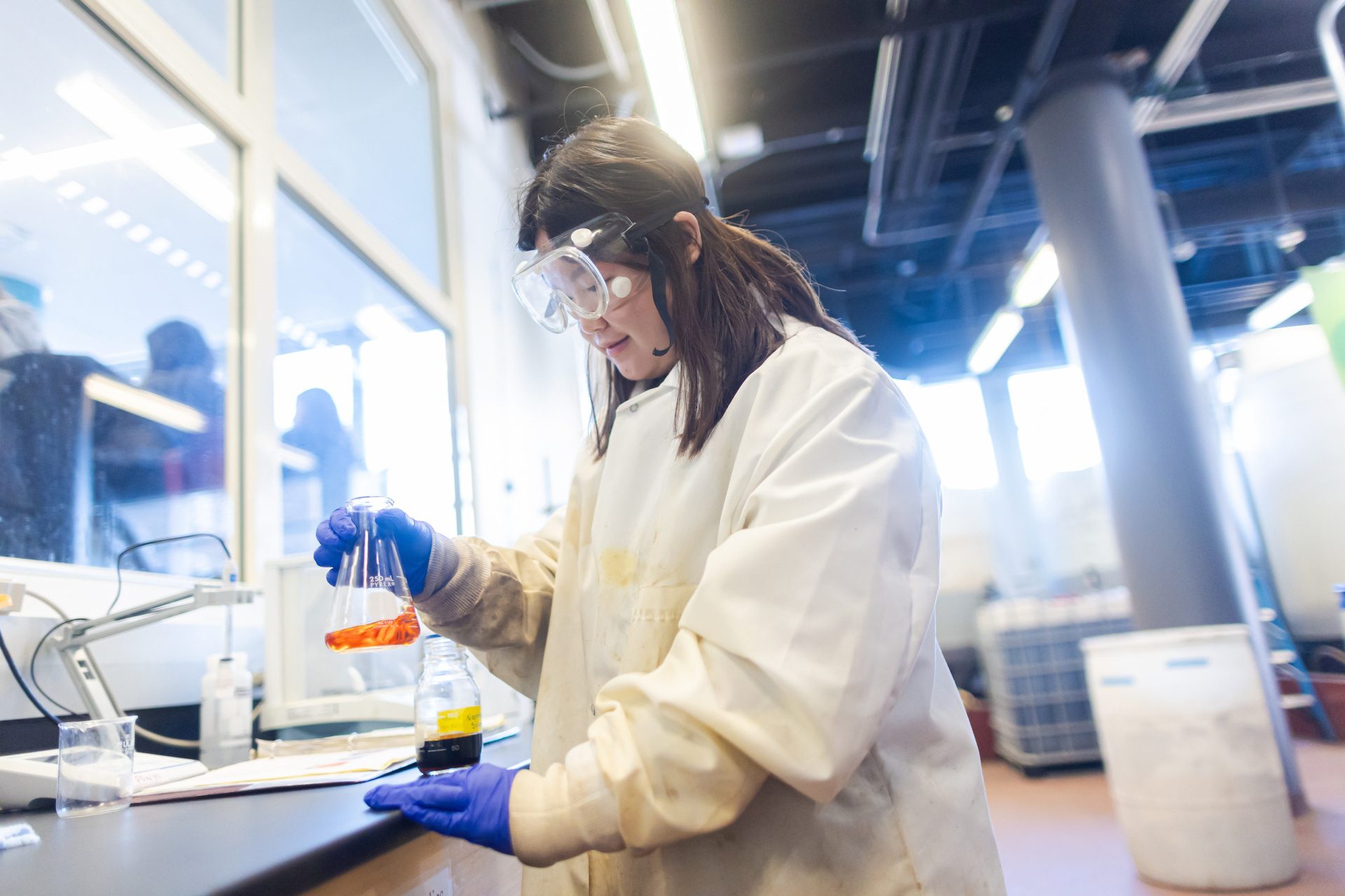 A Loyola University Chicago student wearing a white lab coat, safety goggles, and latex gloves holds a beaker of fluid in a lab on campus