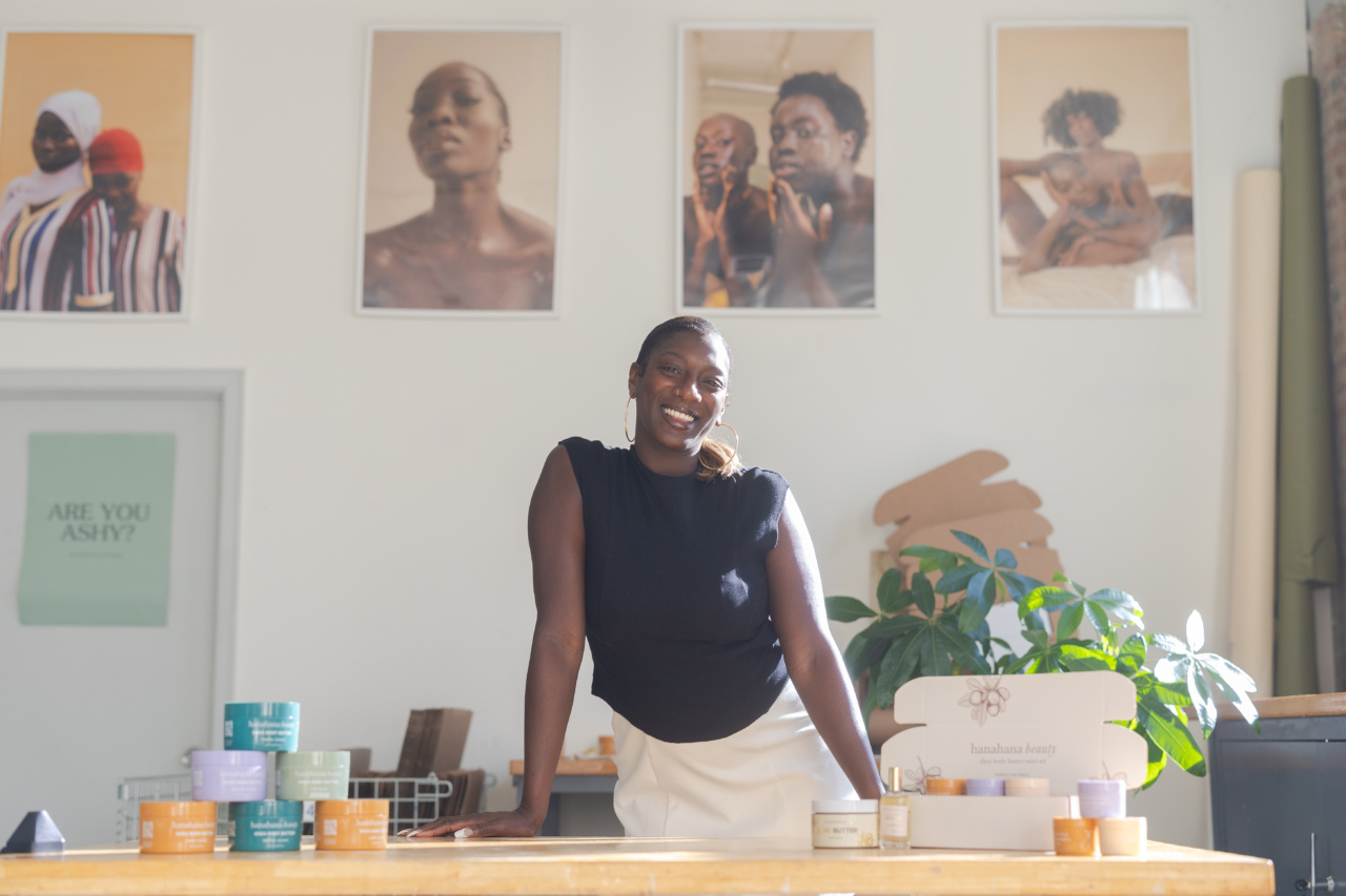 A Black woman poses in front of a desk displaying skincare products with model photos in the background