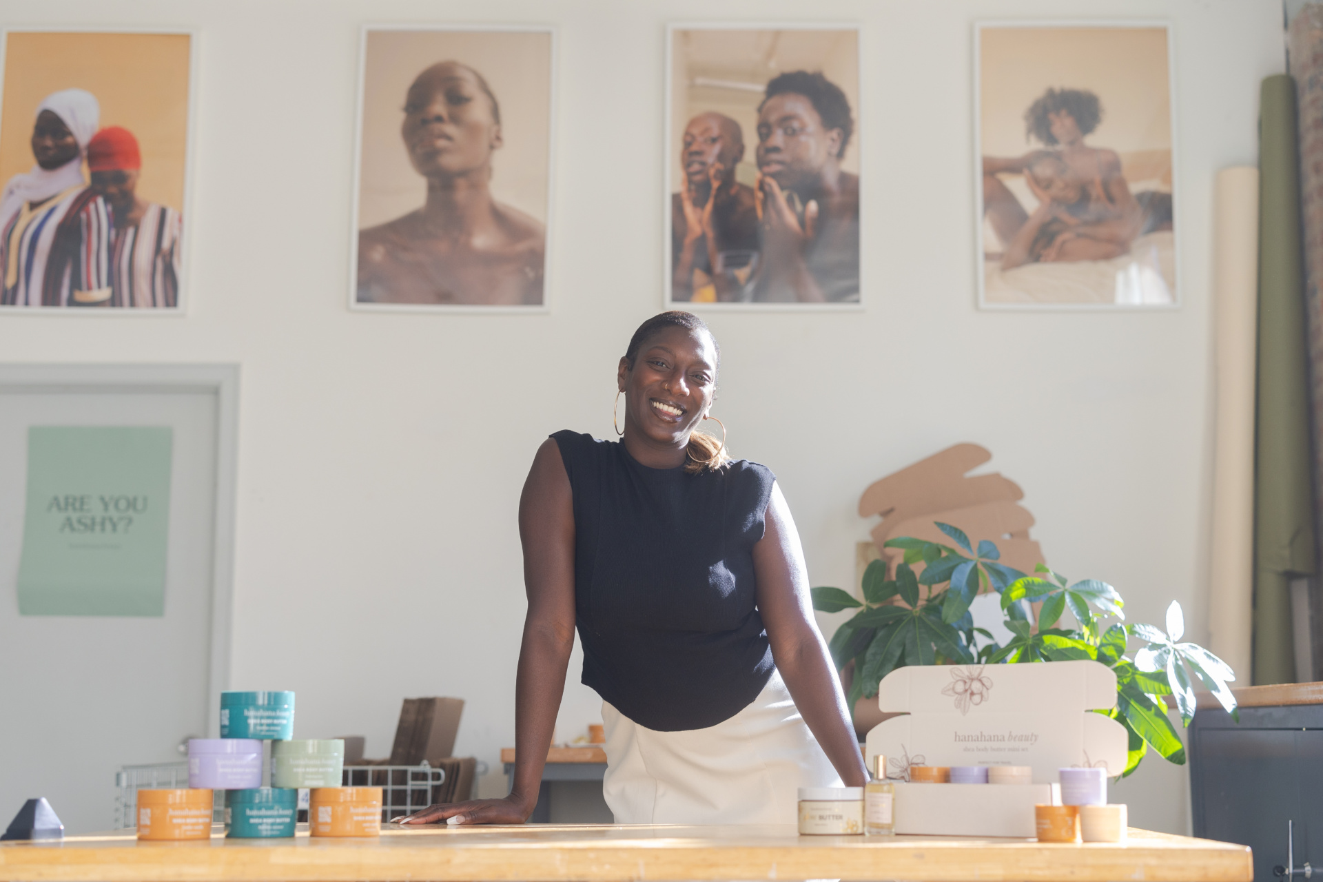 A Black woman poses in front of a desk displaying skincare products with model photos in the background