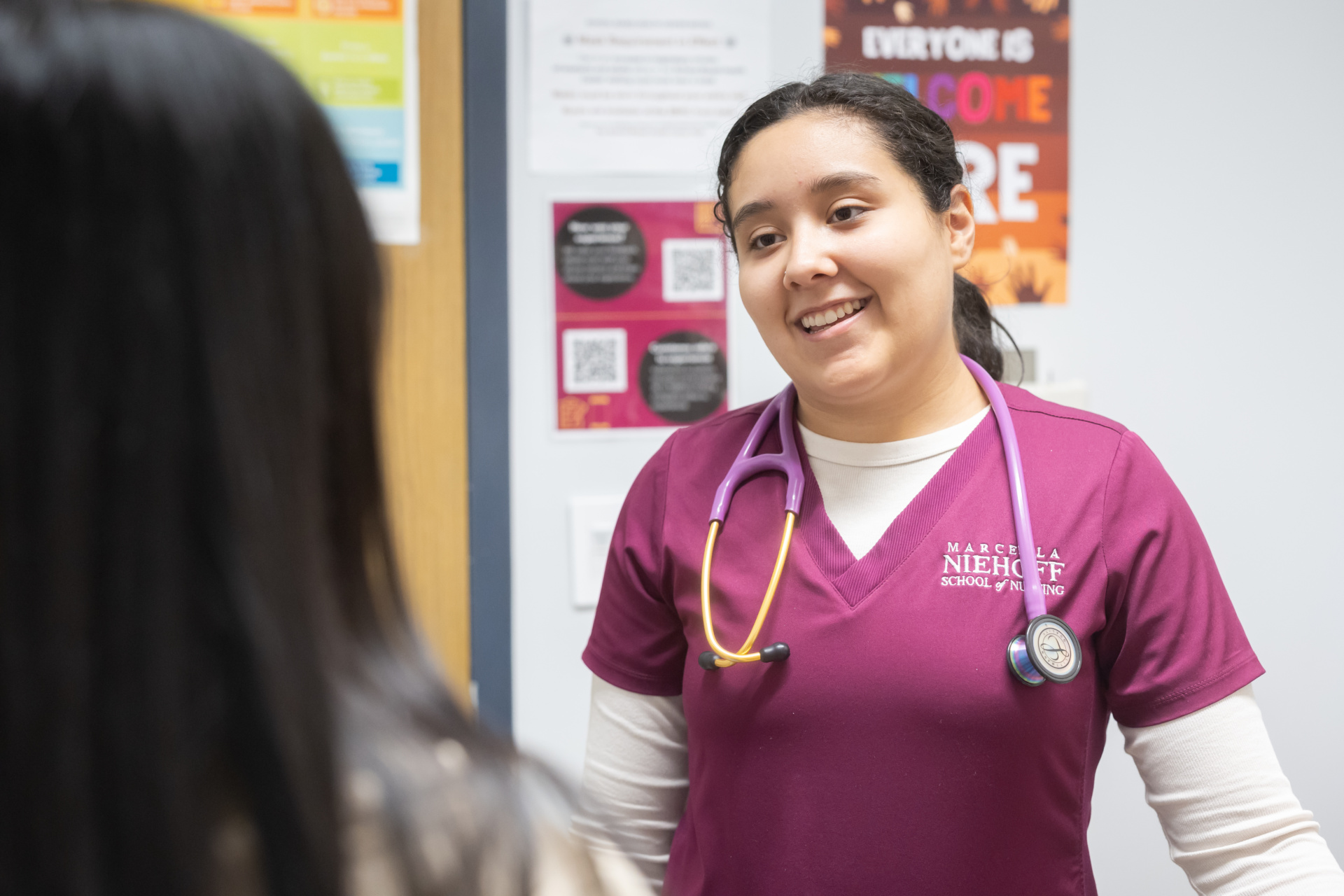 A Loyola University Chicago nursing student wearing scrubs and a stethoscope around her neck speaks to a patient out of frame in a health clinic