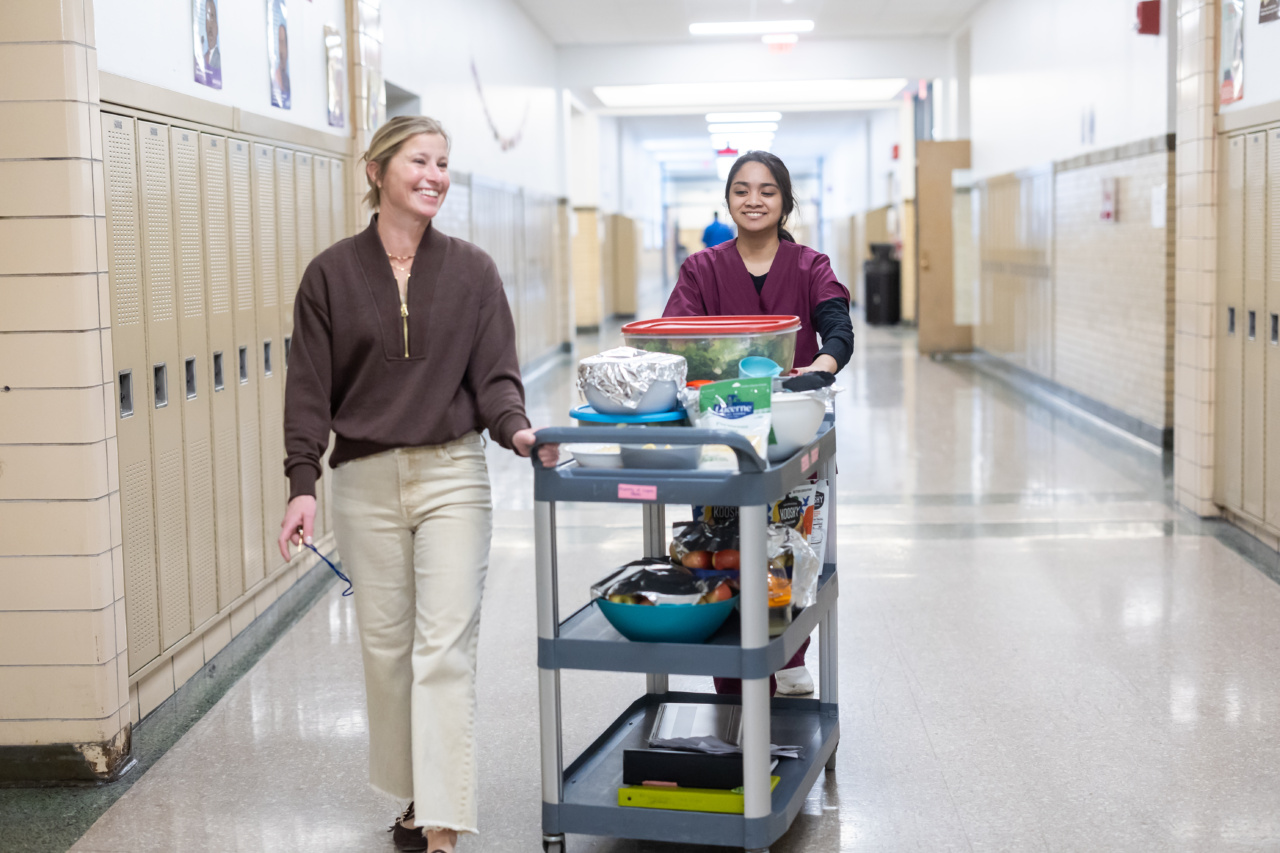 A Loyola University Chicago student pushes a cart of food down a school hallway lined with lockers as a professor walks next to her