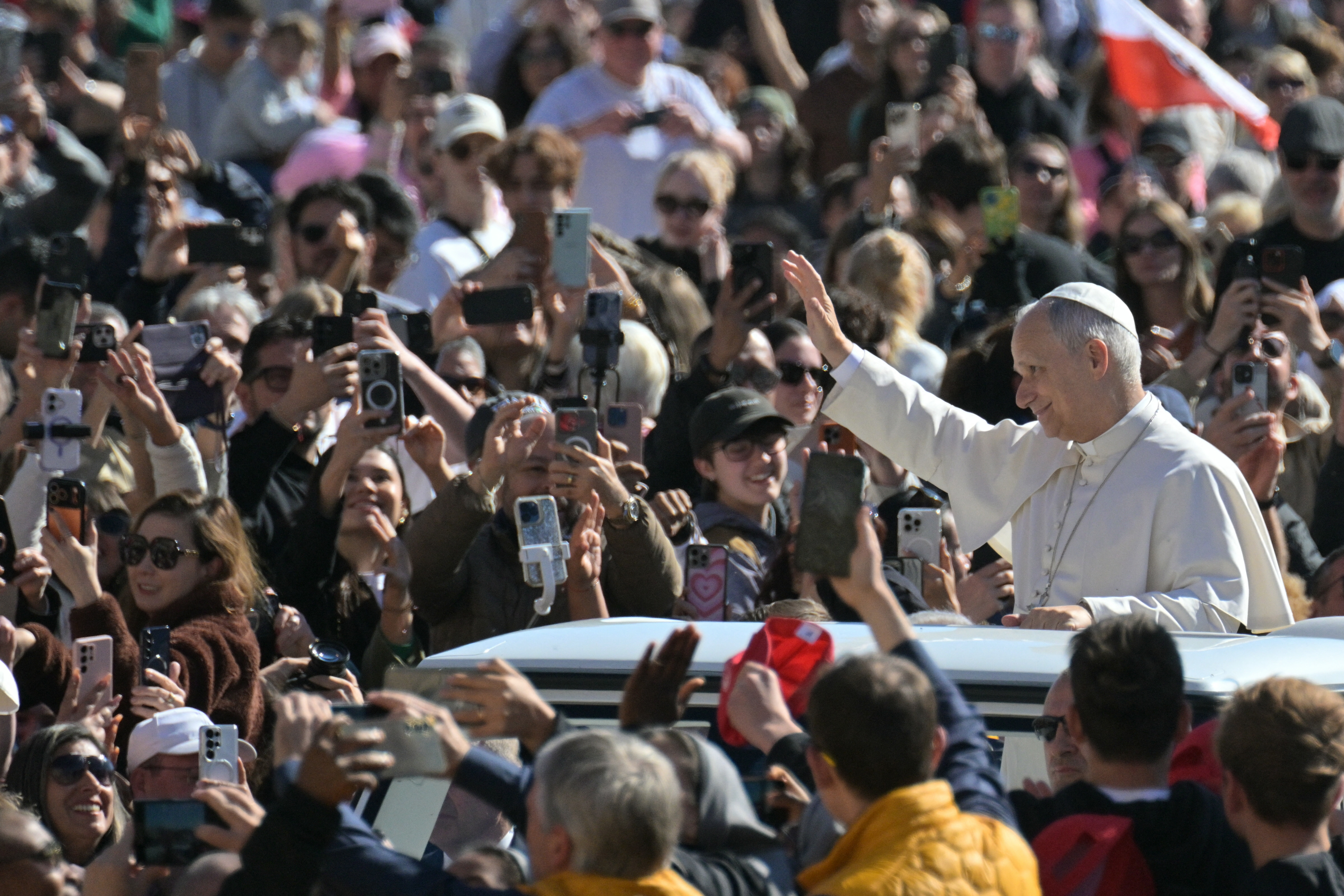 A man dressed in papal white robes stands on a vehicle and waves at a crowd surrounding him and holding up cellphones to record the moment on a sunny day outdoors