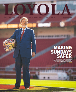 A man wearing a suit stands on a large football field with scarlet stadium seats in the background and holds a football helmet with an 