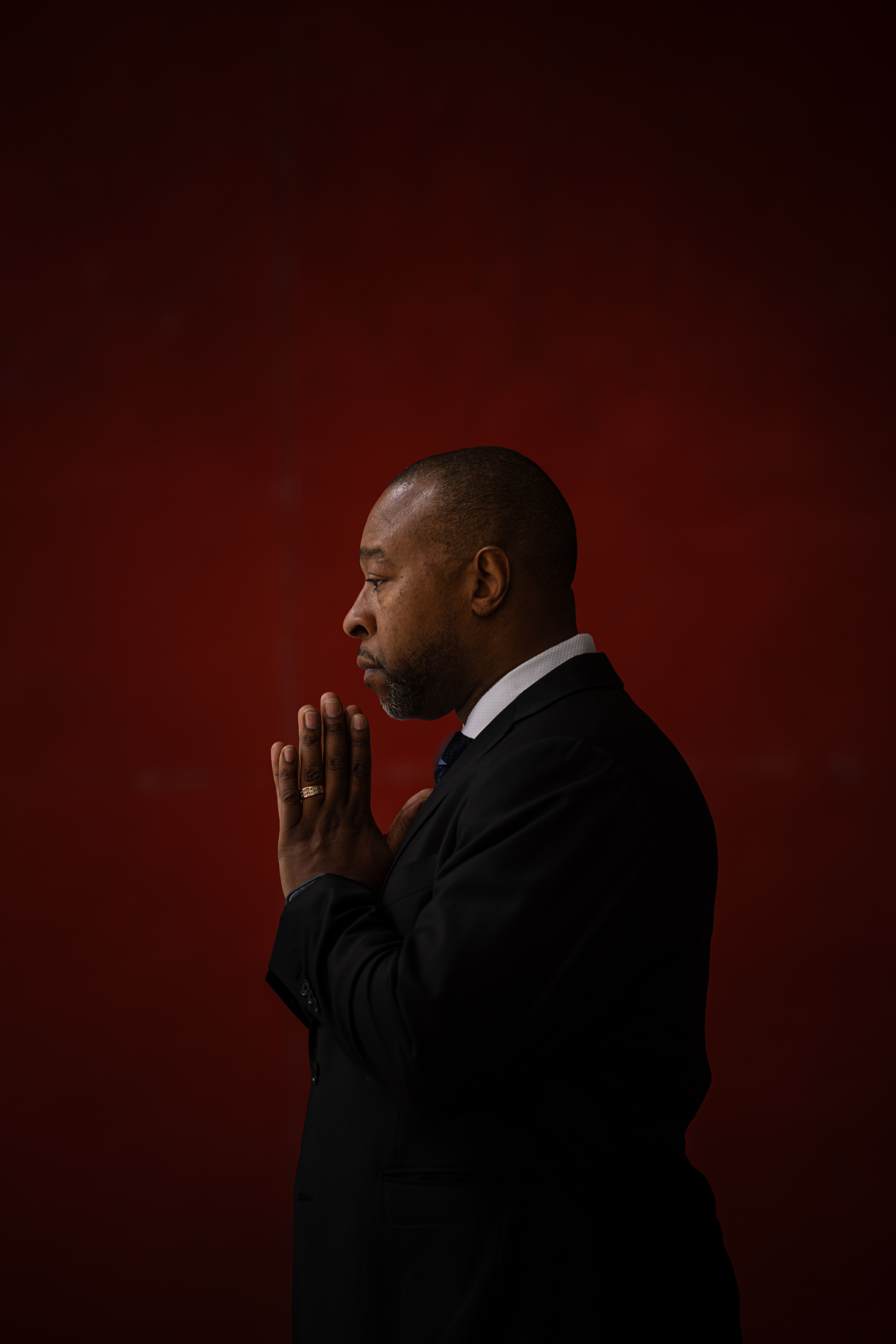 A man in a black suit poses in profile with hands in a prayer position against a red background