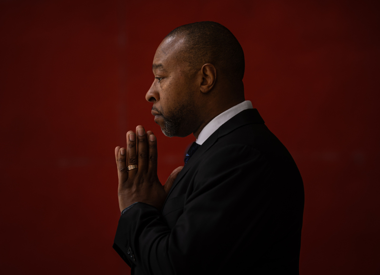 A man in a black suit poses in profile with hands in a prayer position against a red background