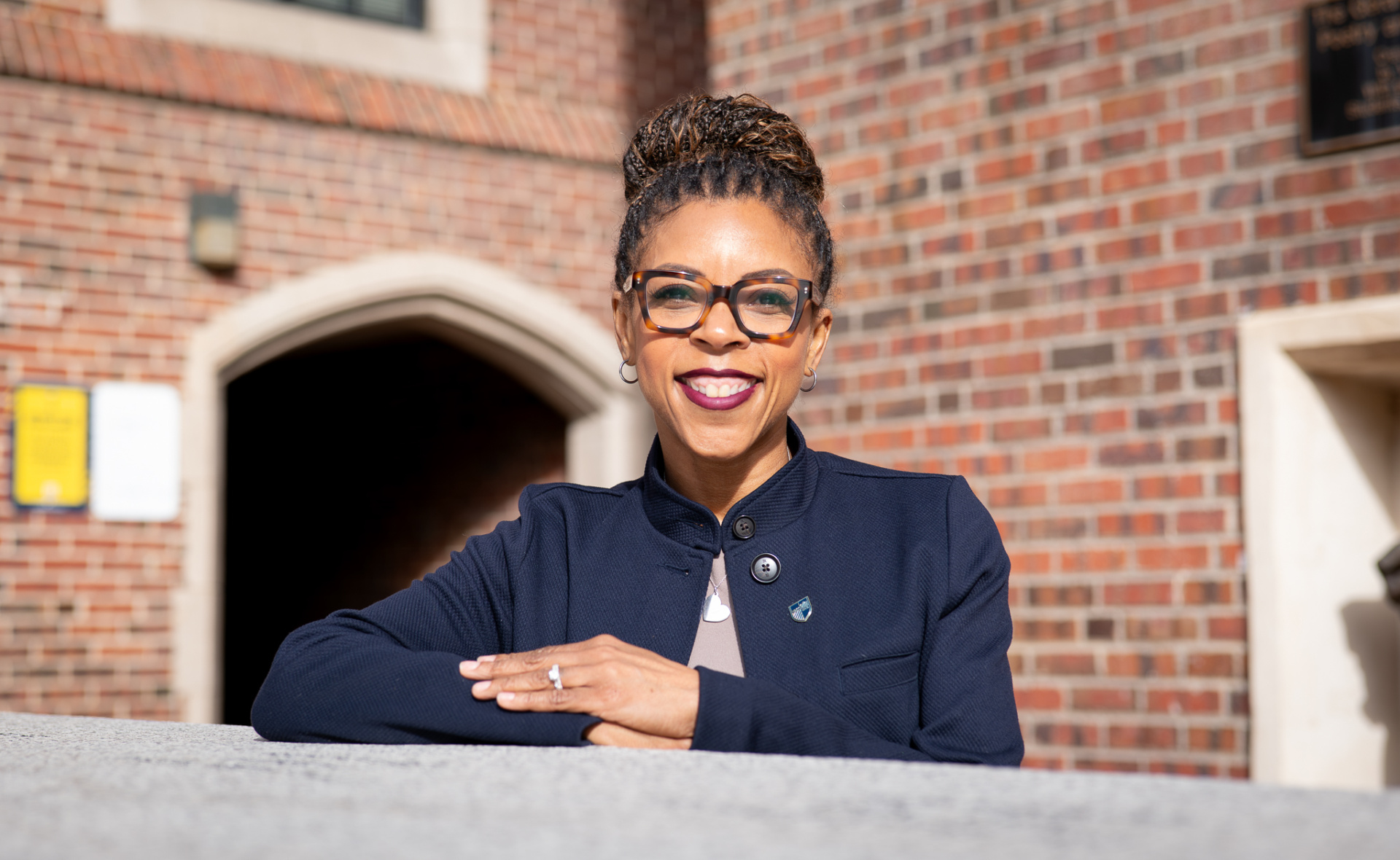 A Black woman wearing glasses and a blue blazer poses outdoors with a brick building in the background