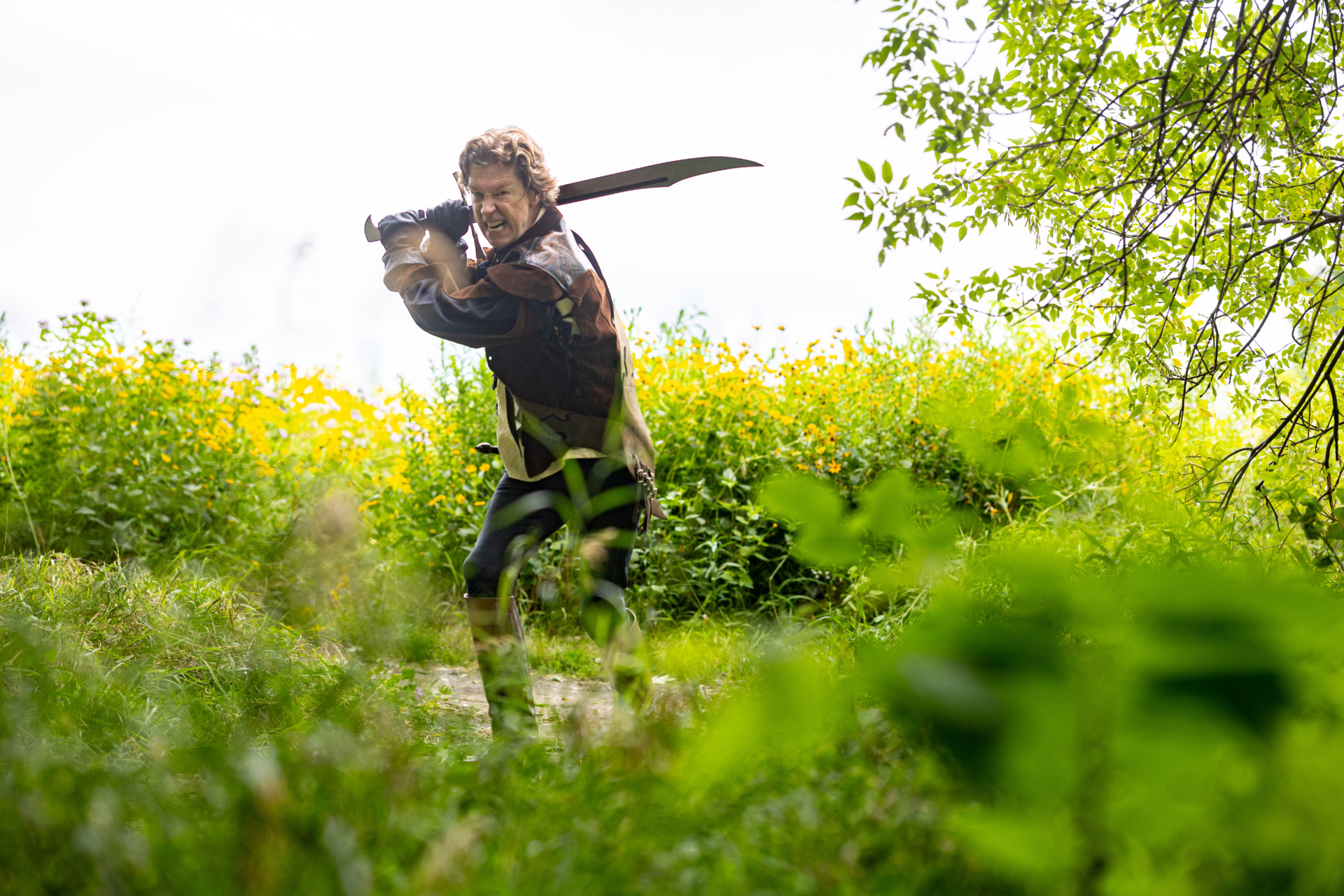 Portrait of Loyola alumnus, Nick Sandys, holding a sword and wearing leather armor outdoors in an overgrown field
