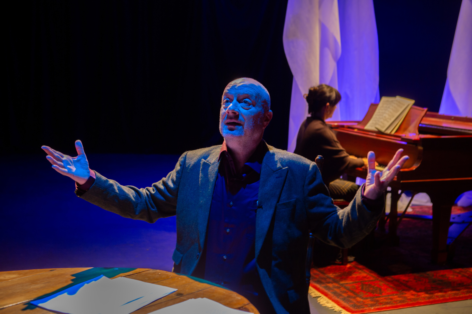 On a theatre stage, an older man in a black suit sits at a wooden table with papers in front of him. Behind him, a woman sits at a piano.