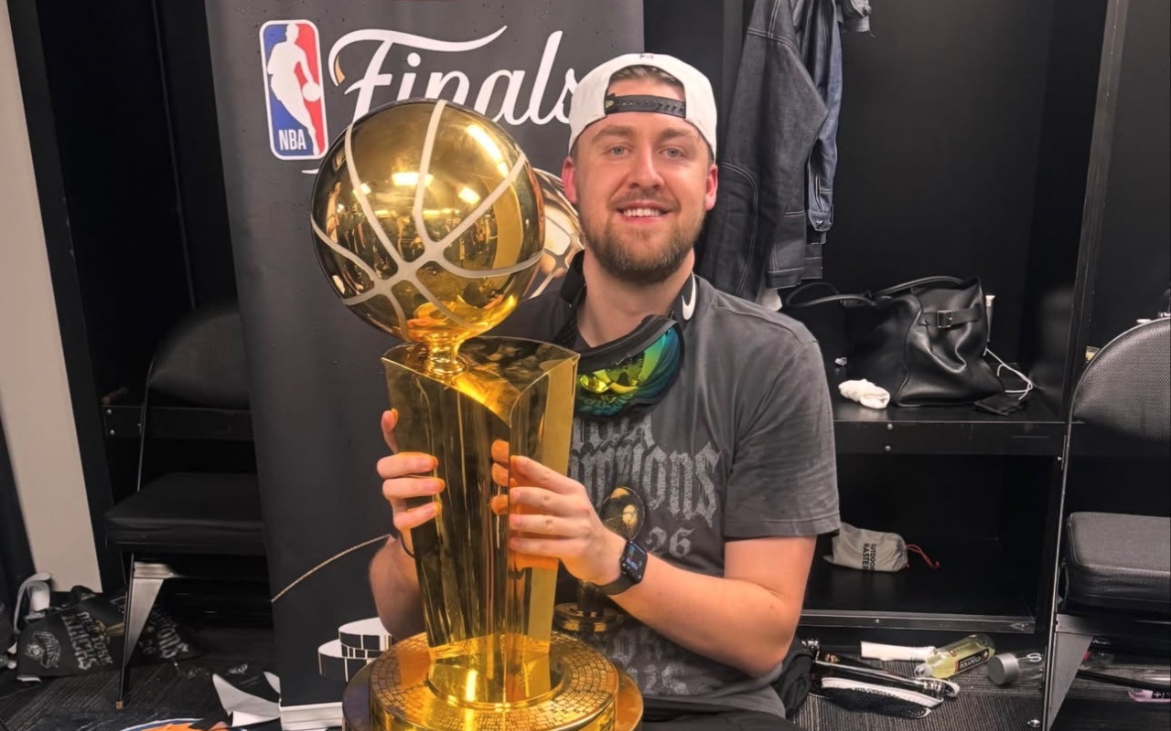 A man wearing a backwards baseball cap holds a large gold trophy while seated in a chair in a locker room